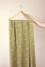 Una mano sujeta una percha de madera de la que cuelga una larga tela de color oliva con flores blancas.