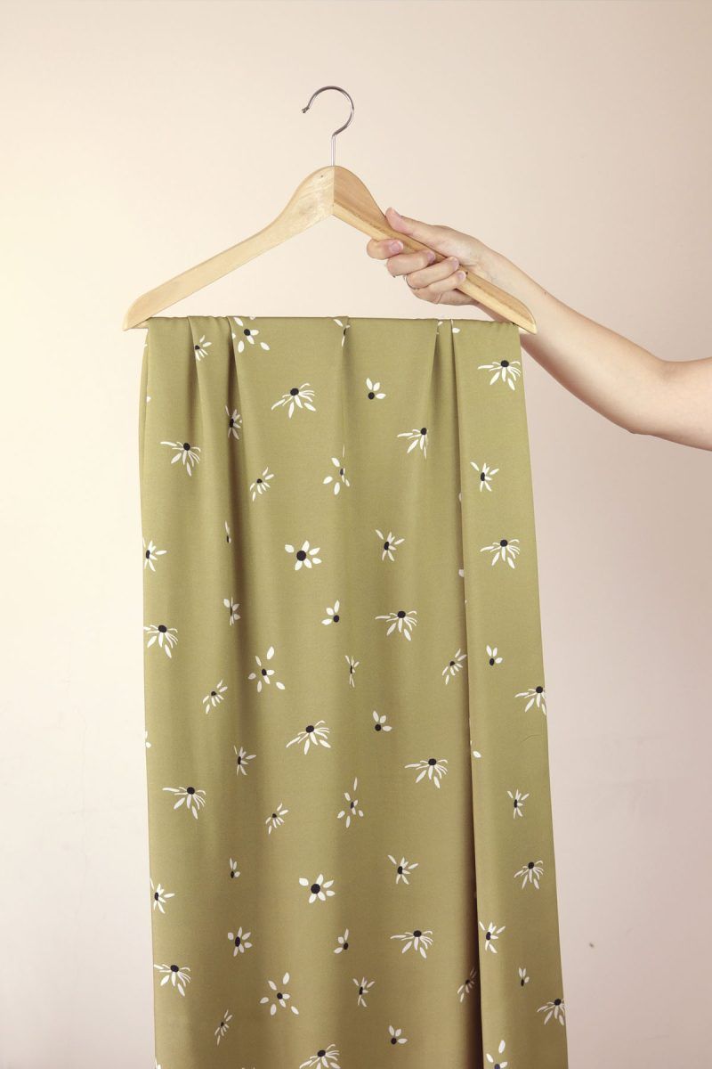 Una mano sujeta una percha de madera de la que cuelga una larga tela de color oliva con flores blancas.