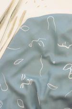 Tejido de viscosa de fondo verde con motivos de caras pictóricas, hojas y líneas simulando el viento en color crema y blanco roto