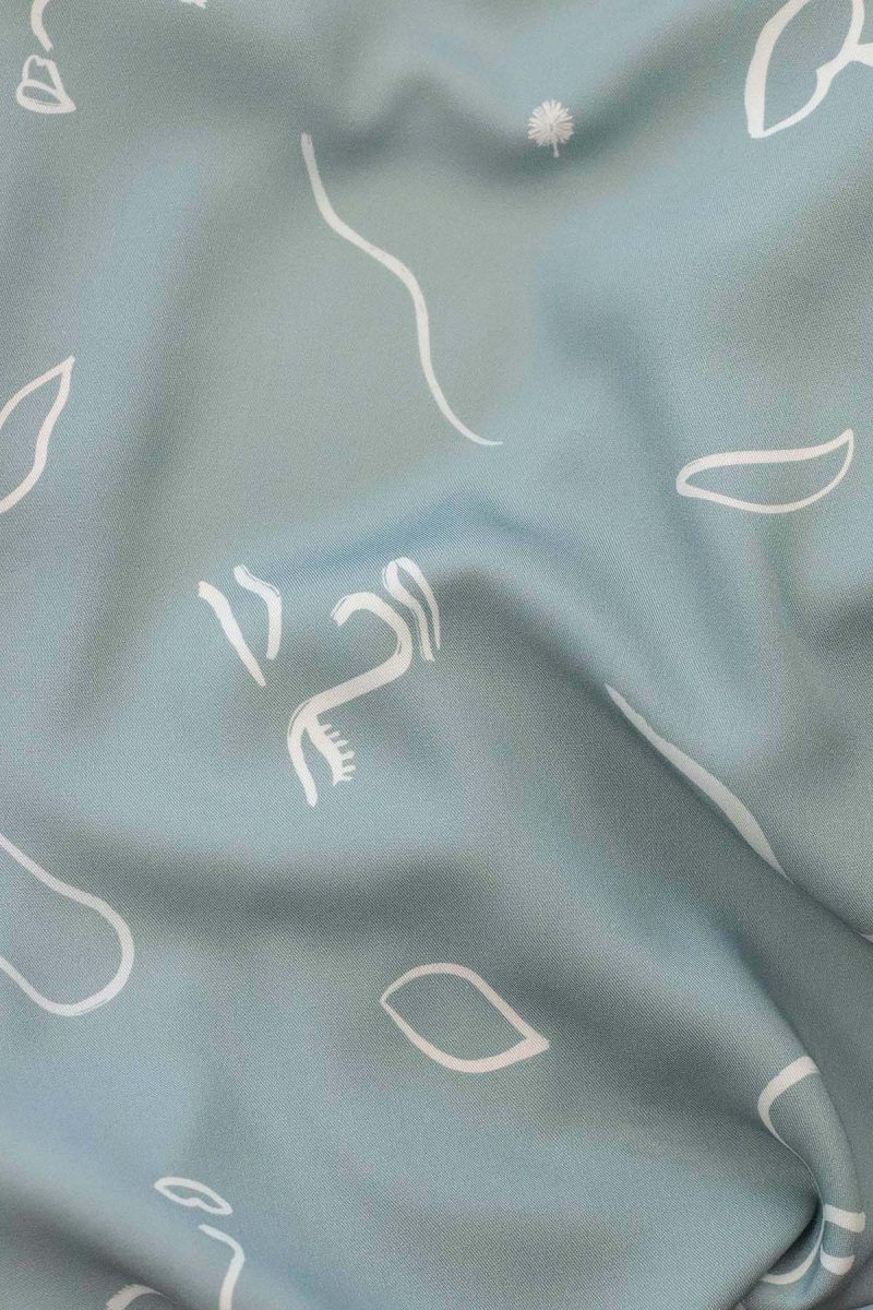 Detalle de tela de viscosa ODALOA con fondo verde con motivos de caras pictóricas, hojas y líneas simulando el viento en color blanco roto y crema