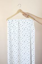 Una mano sujeta una percha de madera de la que cuelga una larga tela de color blanco roto con motivos de pequeñas hojas en colores azules, rosa y marrón.