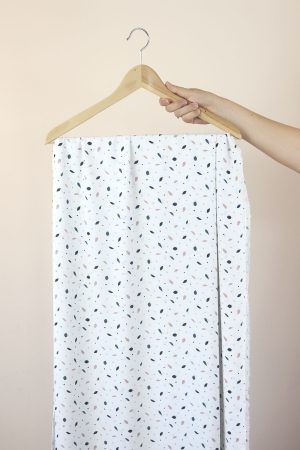 Una mano sujeta una percha de madera de la que cuelga una larga tela de color blanco roto con motivos de pequeñas hojas en colores azules, rosa y marrón.