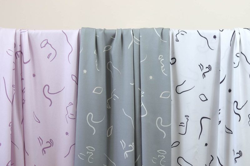 Tres largas telas de viscosa en colores verde grisáceo, malva y blanco roto cuelgan de una barra. Tienen motivos de caras abstractas y líneas de viento.