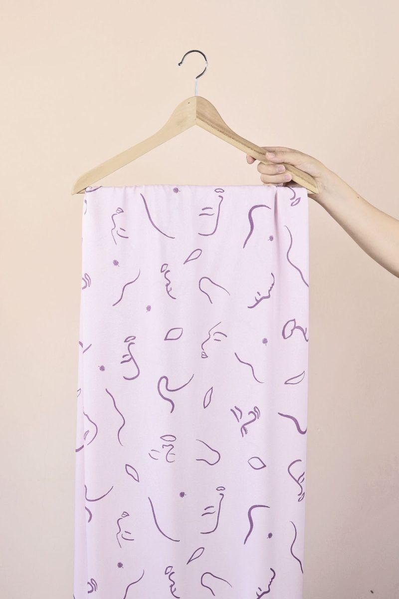 Una mano sujeta una percha de madera de la que cuelga una larga tela de color malva con motivo de caras abstractas, lineas y hojas en malva oscuro.