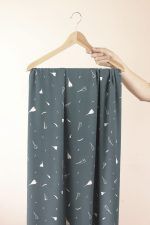 Viscosa lirio de otoño Lago Una mano sujeta una percha de madera de la que cuelga una larga tela de color azul verdoso con flores y pétalos blancos.