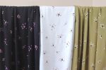 Tres largas telas de viscosa en colores negro, blanco y oliva cuelgan de una barra. Están estampadas con motivos de flores margaritas que empiezan a deshojarse.