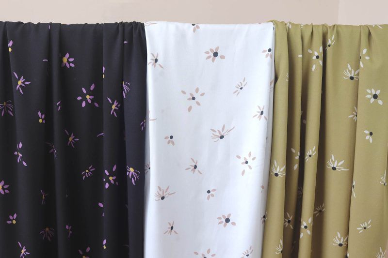 Tres largas telas de viscosa en colores negro, blanco y oliva cuelgan de una barra. Están estampadas con motivos de flores margaritas que empiezan a deshojarse.