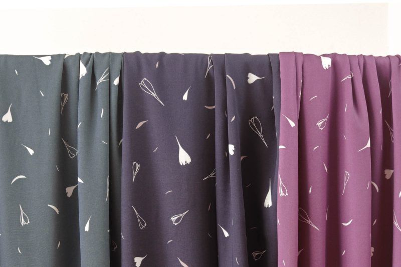 Tres largas telas de viscosa en colores verde azulado, azul oscuro y ciruela cuelgan de una barra. están estampadas con motivos de flores de lirios de otoño y pequeñas hojas en colores blanco.