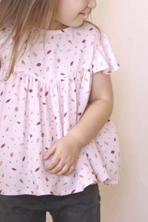 Niña pequeña, de unos tres años, castaña y de pelo largo, lleva una blusa de manga corta, con la viscosa Danza de las Hojas Rosa. La tela es rosa claro con pequeñas hojas en colores rosa más oscuro, burdeos y marrón. Lo combina con unos pantalones vaqueros negros.