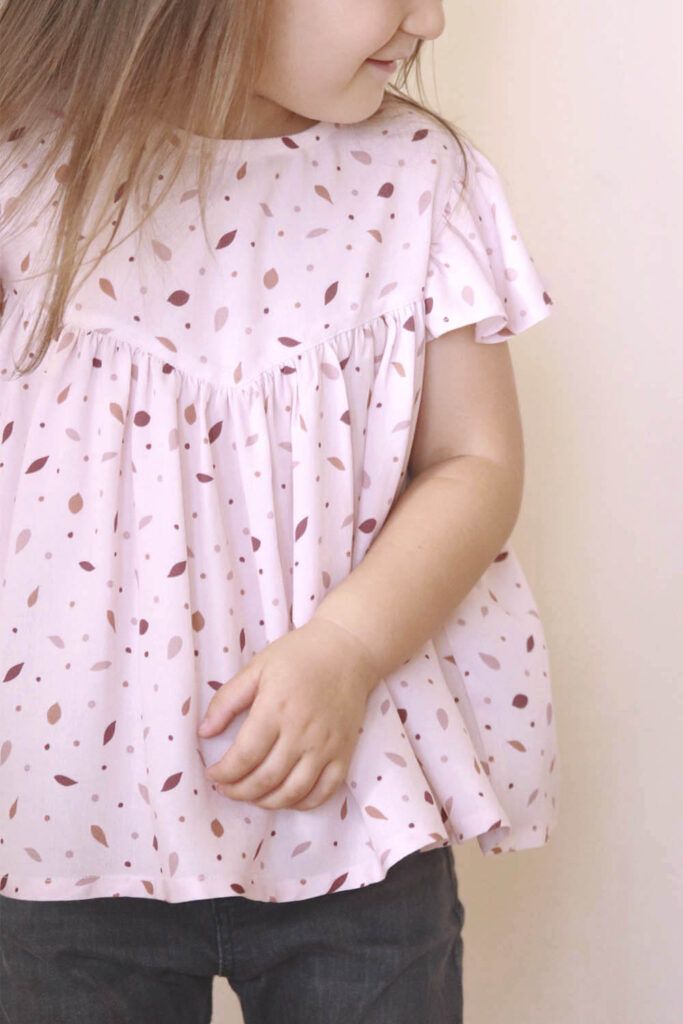 Niña pequeña, de unos tres años, castaña y de pelo largo, lleva una blusa de manga corta, con la viscosa Danza de las Hojas Rosa. La tela es rosa claro con pequeñas hojas en colores rosa más oscuro, burdeos y marrón. Lo combina con unos pantalones vaqueros negros.