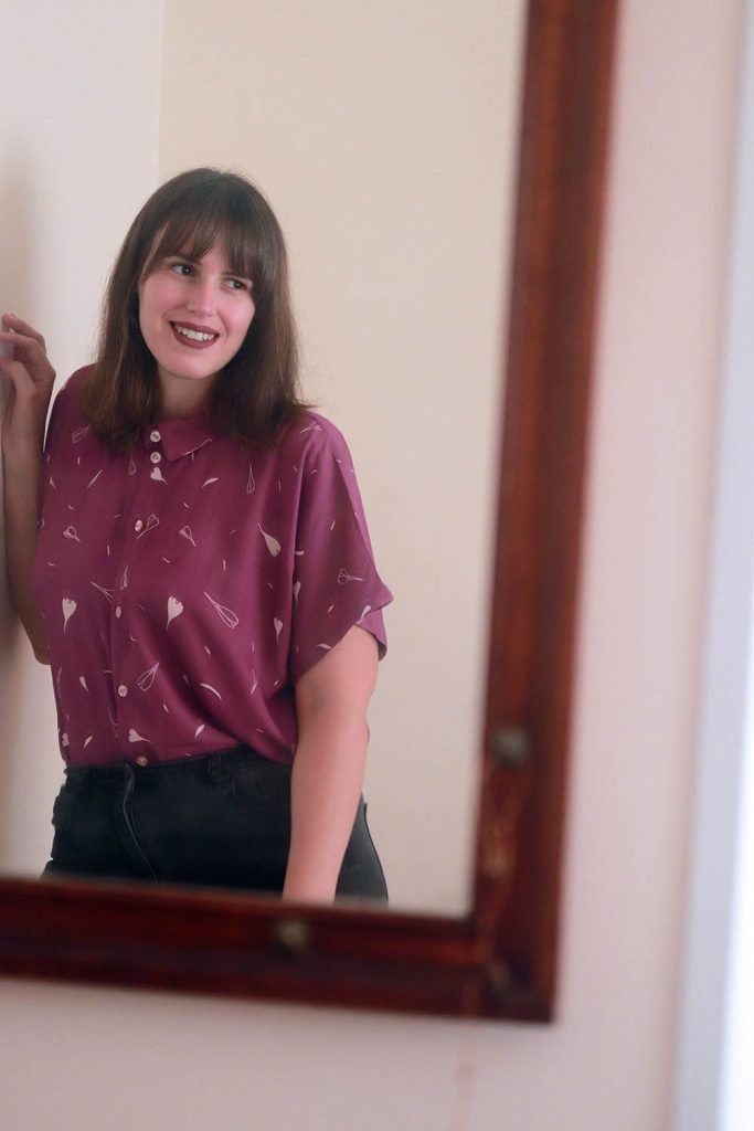 Reflejo en un espejo de una chica con pelo castaño y moreno que lleva una camisa de manga corta con el estampado Lirio de Otoño color ciruela. Este estampado es minimalísta, con flores de lirio de otoño al viento en color blanco.