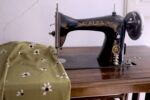 Máquina de coser antigua, negra con letras doradas en la que se puede leer ALFA. Al lado se encuentra una tela de color verde oliva con flores blancas.