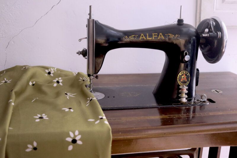 Máquina de coser antigua, negra con letras doradas en la que se puede leer ALFA. Al lado se encuentra una tela de color verde oliva con flores blancas.