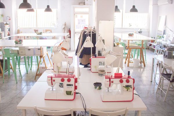 Interior de una academia de costura en la que se ven varias máquinas de coser, mesas altas para hacer patrones y cortar telas, así como un burro con ropa colgada.