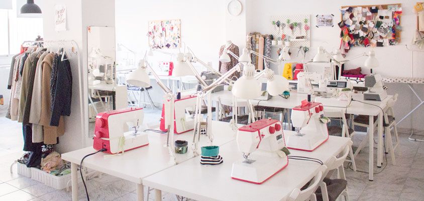 Interior de una academia de costura en la que se ven dos amplias mesas con máquinas de coser. A un lado, un burro perchero con ropa colgando. Al fondo, en la pared, un corcho con muestras de telas e imágenes de inspiración.