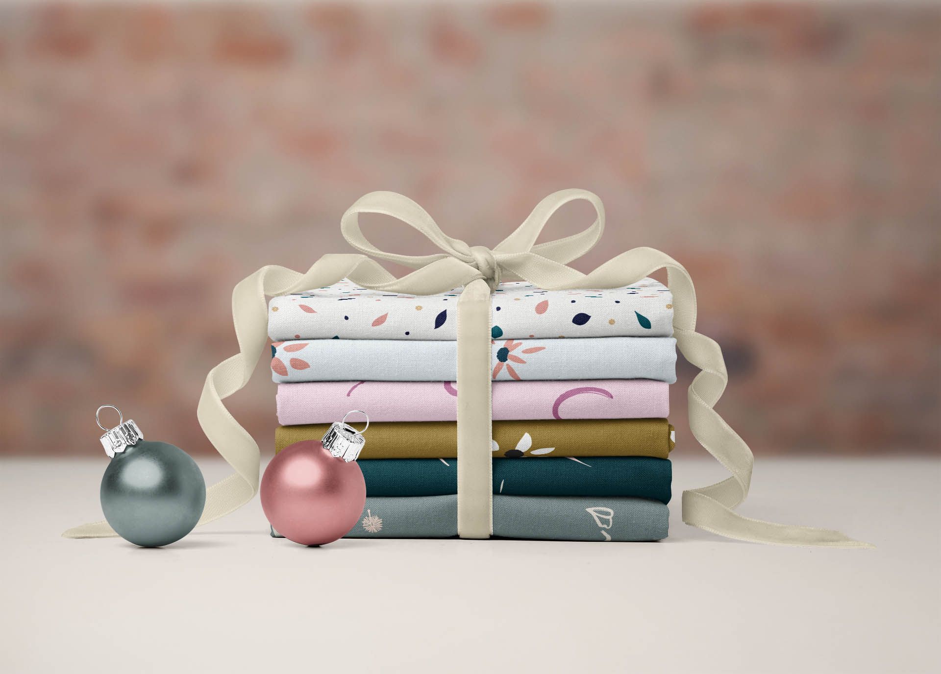 Pack de telas atadas por una cinta de tela, presentado a modo de regalo para costureras. Dos bolas de decoración de árbol de Navidad se encuentran a los pies de las telas.