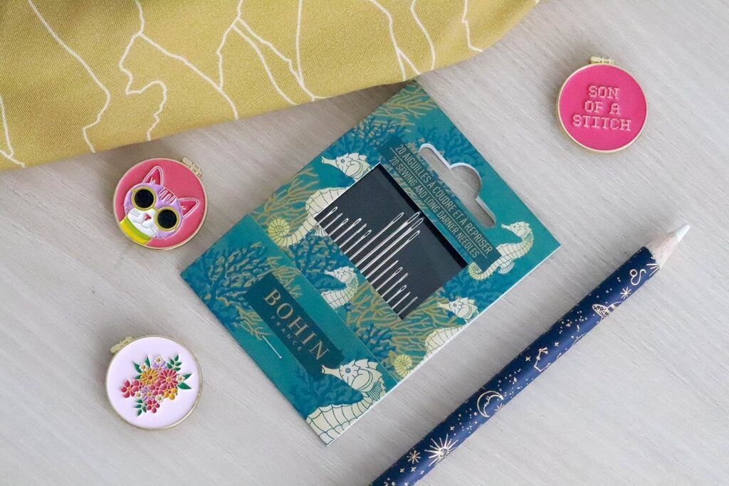 Tres porta agujas imantados con forma de bastidor redondo están encima de una mesa de madera. Uno tiene una cara de gatito, otro una ramo de flores y en otro se lee "son of a stitch". A su lado se encuentra un pack de agujas con packing de caballitos de mar y corales, un lápiz de tiza para marcar la tela con motivos de constelaciones y una tela color mostaza con líneas blancas que forman montañas.