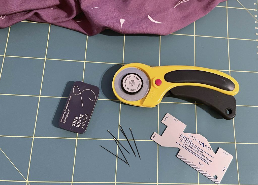 Sobre una base de corte se encuentra un cúter rotatorio, un puntímetro, una caja de alfileres y alfileres sueltos y una tela color ciruela.