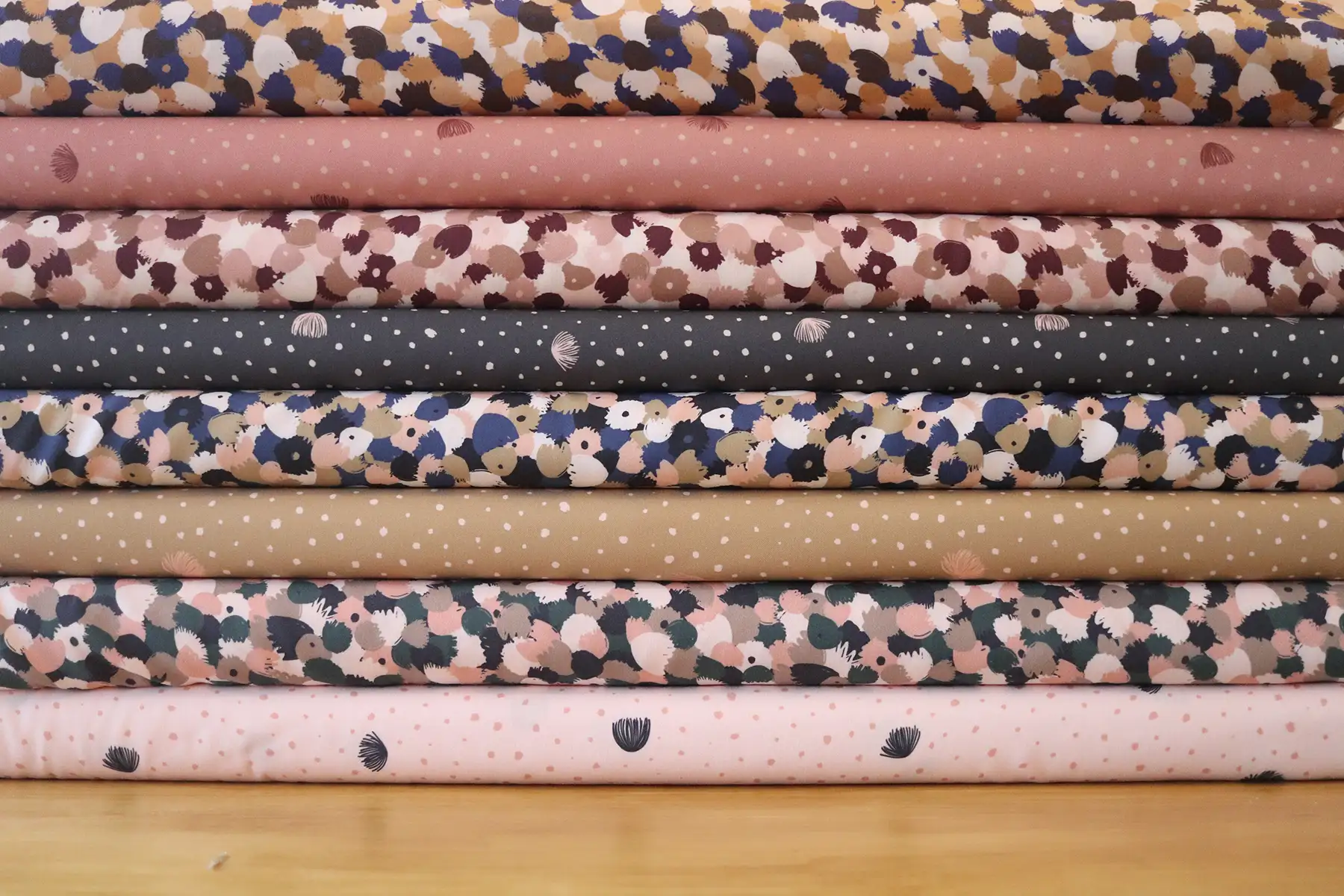 Ocho tablillas de telas, apiladas una encima de otra, sobre una mesa marrón. Cuatro de estas telas son de flores abstractas en colores azules, rosados, verdes, blancos y marrones. Las otras cuatro tienen estampados de puntitos con flores del árbol de la seda. Tienen colores salmón, ocre, rosado y gris oscuro.