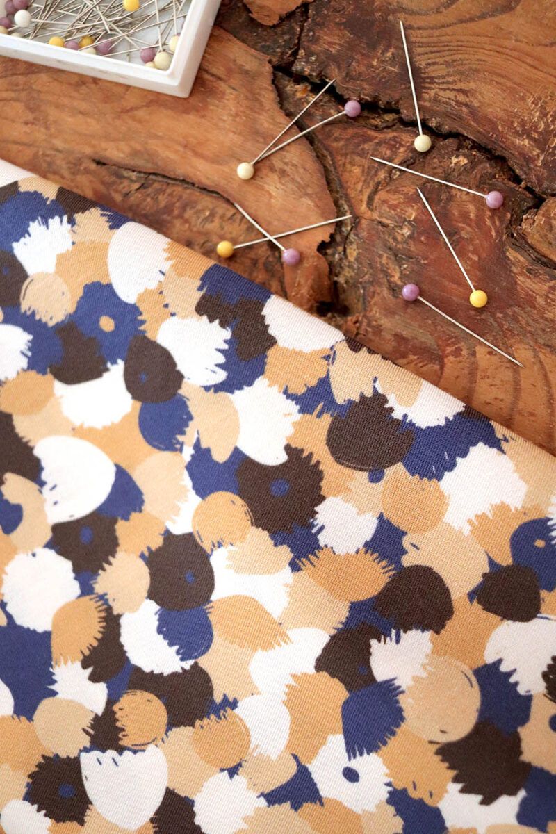 Viscosa sostenible_Mille Fiori_Desierto Tela de flores pequeñas y abstractas en color azul marino, azul intenso, marrón arena y blanco sobre un trozo de madera rústico. Al lado de la tela se puede observar algunos alfileres con cabeza bola, unos en tono amarillo, otros lila y otros blancos.