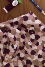 Tela de flores pequeñas en color burgundy, marrón, diferentes tonos de rosa palo y blanco crudo. Está sobre una tabla de madera. A su lado se encuentra un carrete de hilo en color burgundy y un lápiz de tiza para marcar la tela.