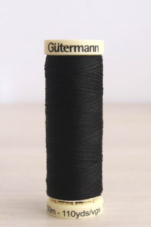 Bobina de hilo Gütermann negro, sobre una madera de color claro y de fondo una pared blanca.
