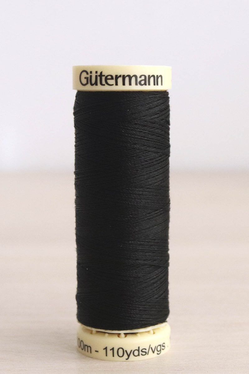 Hilo Gütermann poliéster color negro nº 000 Bobina de hilo Gütermann negro, sobre una madera de color claro y de fondo una pared blanca.