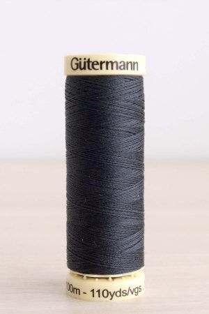 Bobina de hilo Gütermann 095, de color gris oscuro, sobre una madera de color claro y de fondo una pared blanca.