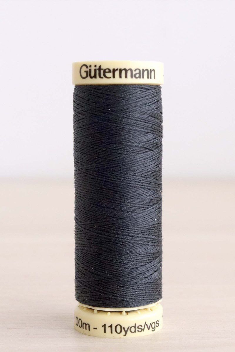 Bobina de hilo Gütermann 095, de color gris oscuro, sobre una madera de color claro y de fondo una pared blanca.