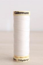Bobina de hilo Gütermann 111, de color blanco crudo, sobre una madera de color claro y de fondo una pared blanca.