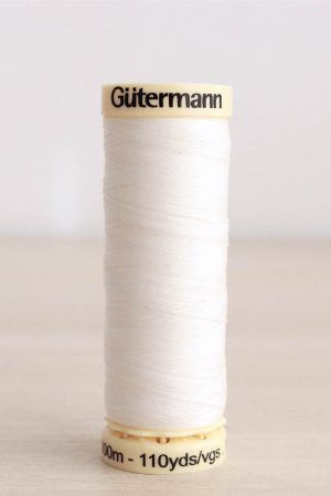 Bobina de hilo Gütermann 111, de color blanco crudo, sobre una madera de color claro y de fondo una pared blanca.