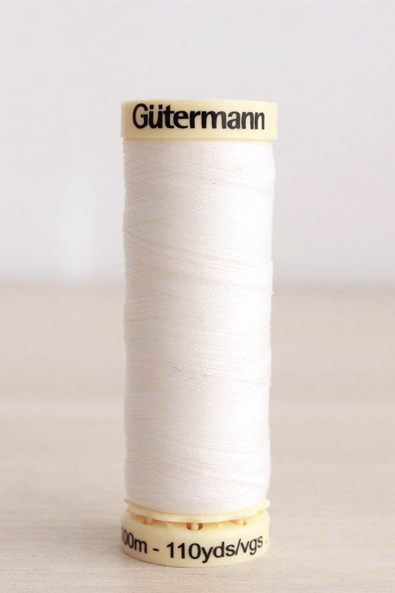 Bobina de hilo Gütermann 111, de color blanco crudo, sobre una madera de color claro y de fondo una pared blanca.