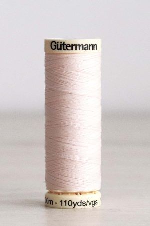 Bobina de hilo Gütermann 210, de color salmón claro, sobre una madera de color claro y de fondo una pared blanca.