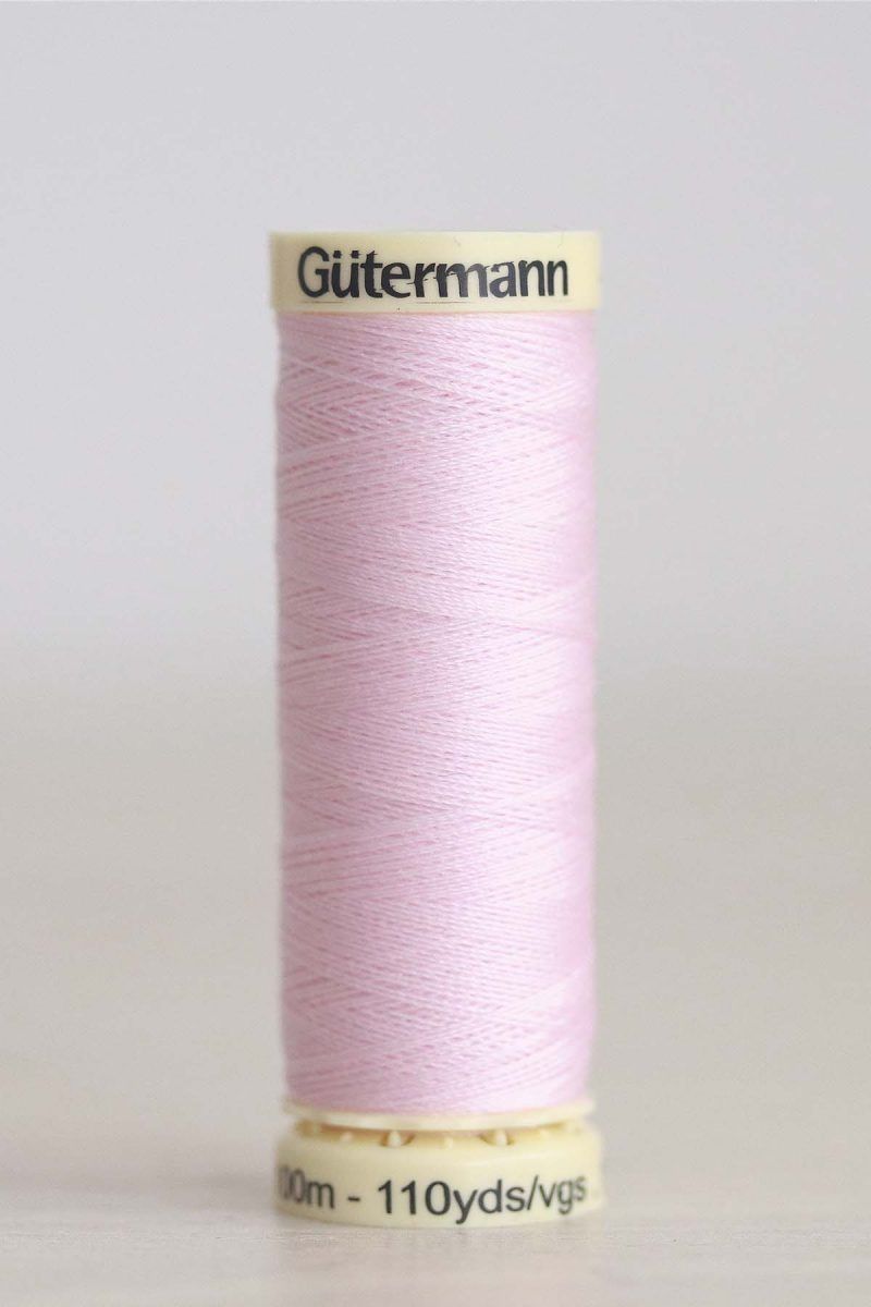 Hilo Gütermann poliéster nº 372 Bobina de hilo Gütermann 372, de color rosa malva, sobre una madera de color claro y de fondo una pared blanca.