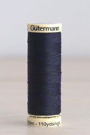 Bobina de hilo Gütermann 387, de color azul oscuro, sobre una madera de color claro y de fondo una pared blanca.