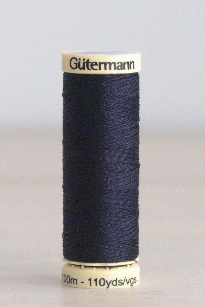 Bobina de hilo Gütermann 387, de color azul oscuro, sobre una madera de color claro y de fondo una pared blanca.