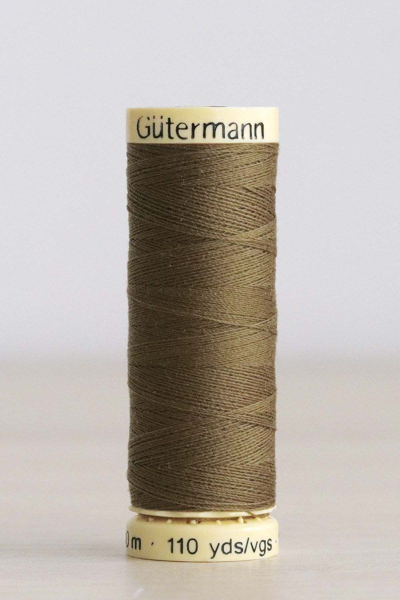 Bobina de hilo Gütermann 528, de color oliva, sobre una madera de color claro y de fondo una pared blanca.