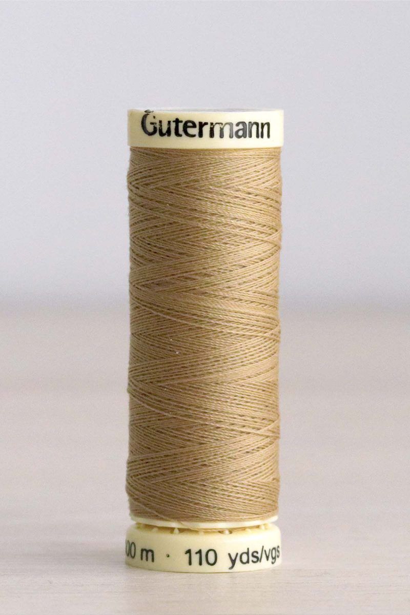 Hilo Gütermann poliéster nº 591 Bobina de hilo Gütermann 591, de color ocre, sobre una madera de color claro y de fondo una pared blanca.