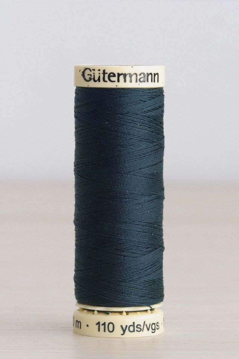 Hilo Gütermann poliéster nº 596 Bobina de hilo Gütermann 596, de color azul verdoso muy oscuro, sobre una madera de color claro y de fondo una pared blanca.