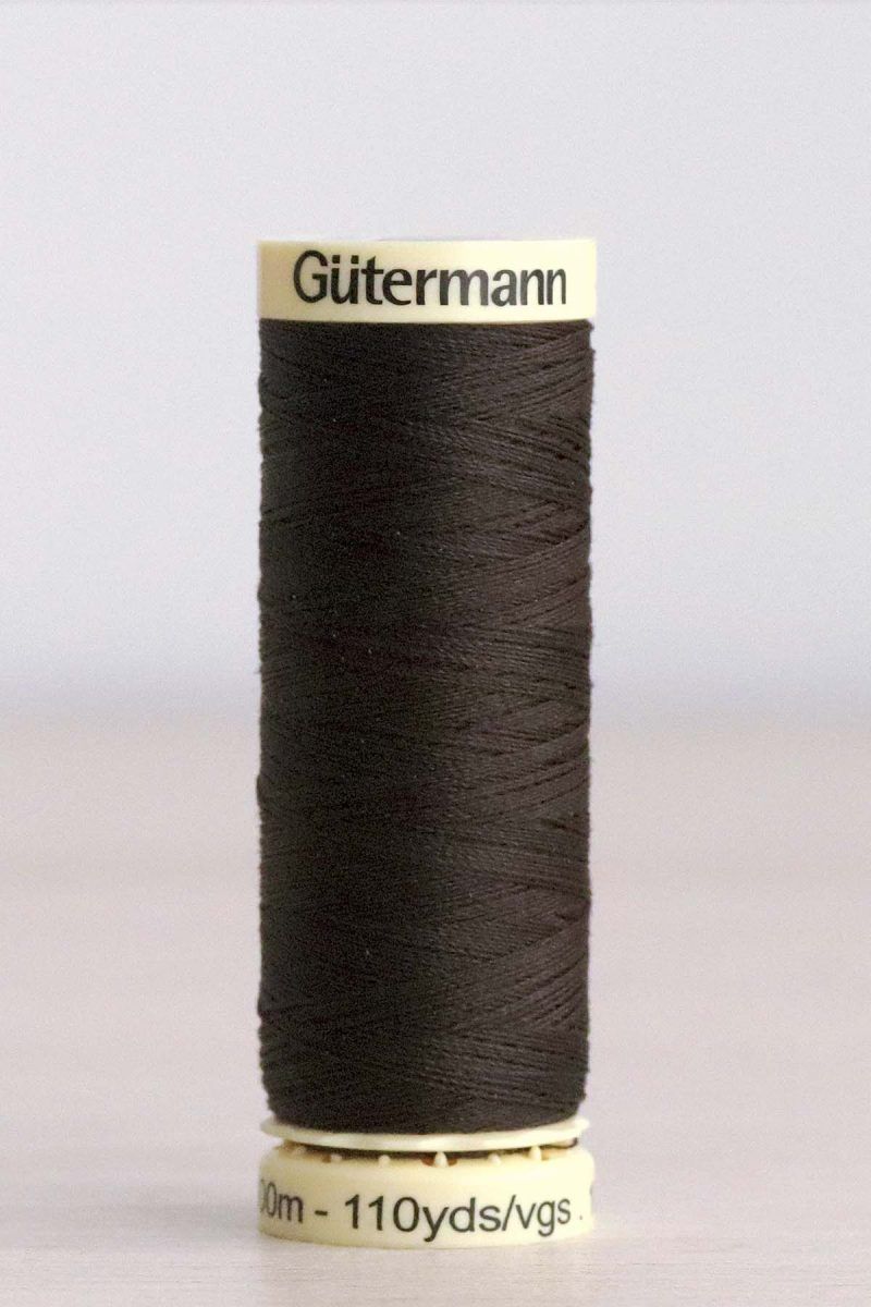 Bobina de hilo Gütermann 697, de color marrón muy oscuro, sobre una madera de color claro y de fondo una pared blanca.