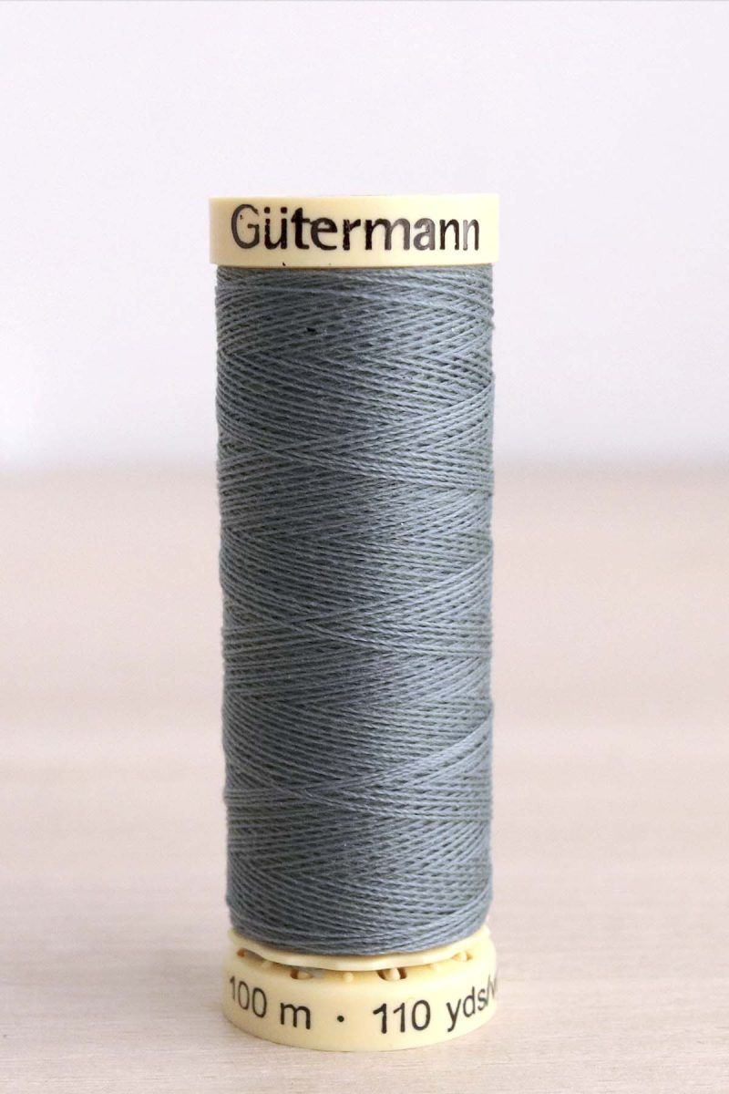 Hilo Gütermann poliéster nº 788 Bobina de hilo Gütermann 788, de color gris verdoso sobre una madera de color claro y de fondo una pared blanca.