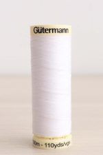 Bobina de hilo Gütermann 800, de color blanco puro, sobre una madera de color claro y de fondo una pared blanca.