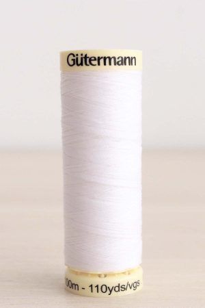 Bobina de hilo Gütermann 800, de color blanco puro, sobre una madera de color claro y de fondo una pared blanca.