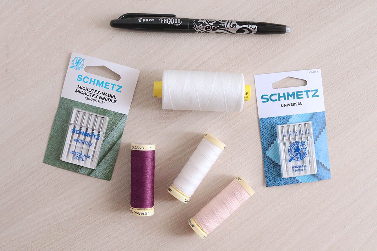 Sobre una mesa se encuentran tres bobinas de hilo pequeñas en color ciruela, blanco crudo y color salmón, una bobina de hilo grande en color blanco crudo, dos blisters de agujas para máquina de coser y un bolígrafo negro Pilot Frixion.