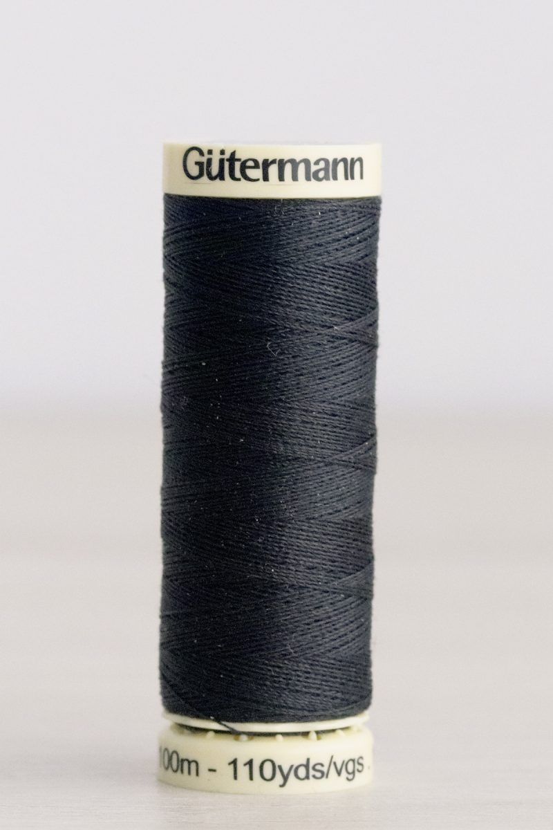Bobina de hilo Gütermann 542, de color gris muy oscuro, sobre una madera de color claro y de fondo una pared blanca.