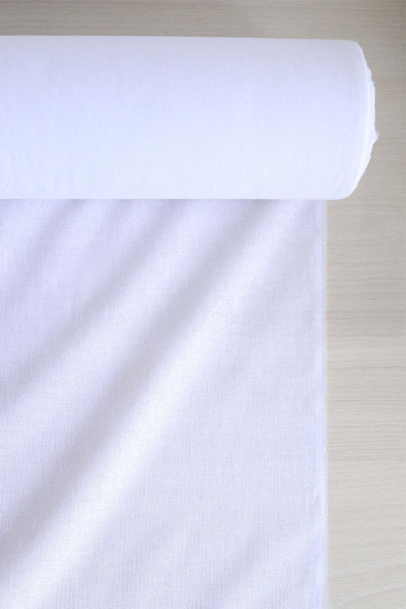 Rollo de entretela termoadhesiva de color blanco sobre una mesa de madera. Una parte de la entretela está desenrrollada y se encuentra estirada sobre la mesa. Esta parte brilla al ser la cara con pegamento.