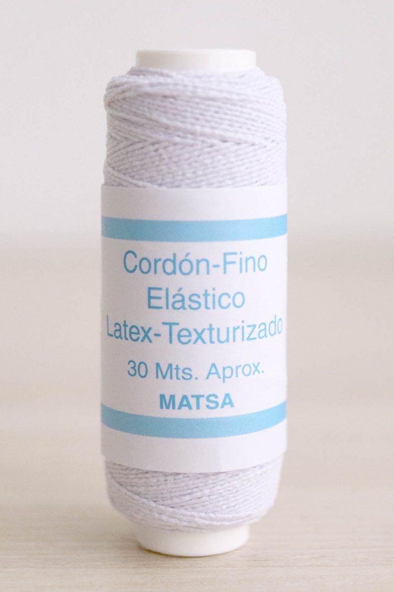 Bobina de hilo elástico para fruncir de color blanco sobre mesa de madera y de fondo una pared blanca.