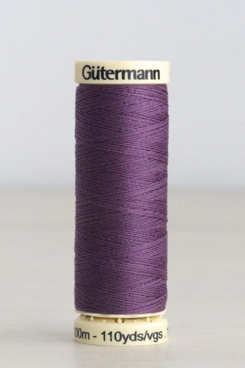 Hilo Gütermann poliéster nº 120 Bobina de hilo Gütermann 120, de color morado, sobre una madera de color claro y de fondo una pared blanca.