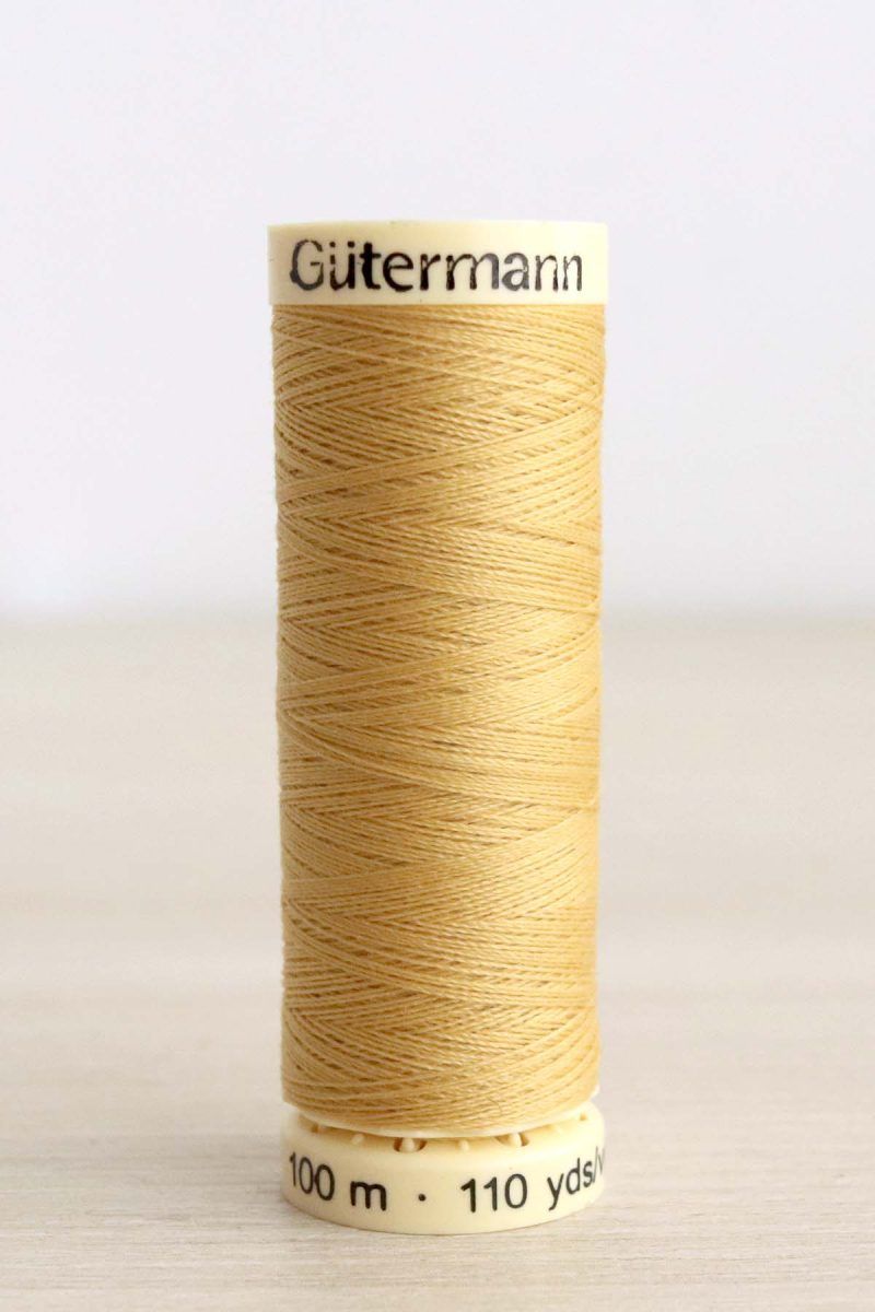 Bobina de hilo Gütermann 488, de color amarillo mostaza, sobre una madera de color claro y de fondo una pared blanca.