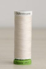 Bobina de hilo Gütermann 135 rPET, de blanco crema, sobre una madera de color claro y de fondo una pared blanca.